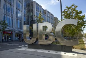 Centred in the image are three large, shiny steel letters reading UBC. Behind them are a row of trees, with a street on either side.