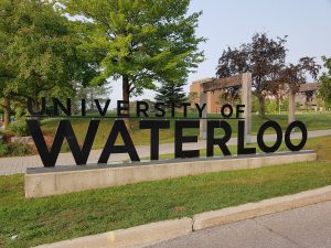 Large letters are cut out of black metal to spell University of Waterloo. Behind the sign is a sidewalk through a grassy area.