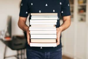 A person wearing a dark blue shirt and jeans holds a stack of books. They’re head and lower body is out of the shot.