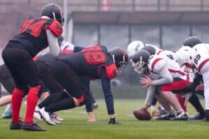 Two football teams are in the snap ahead of open play. Three players in red and black jersey's face the opposing team's center holding the ball, wearing red and white jerseys.